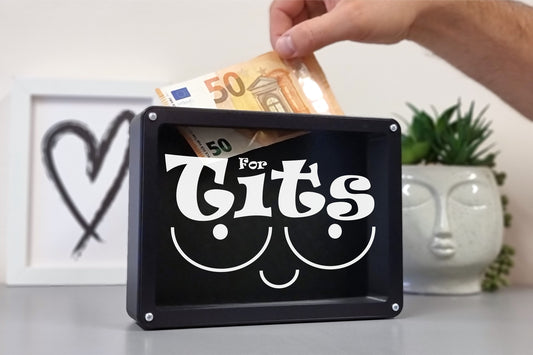 A humorous black money box featuring the word "Tits" in a playful design, with a hand inserting a 50-euro note.