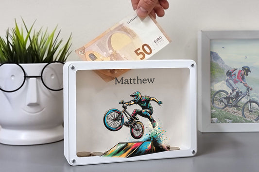 A white personalized money box featuring a motocross bike design with the name "Matthew" and a hand inserting a 50-euro note.